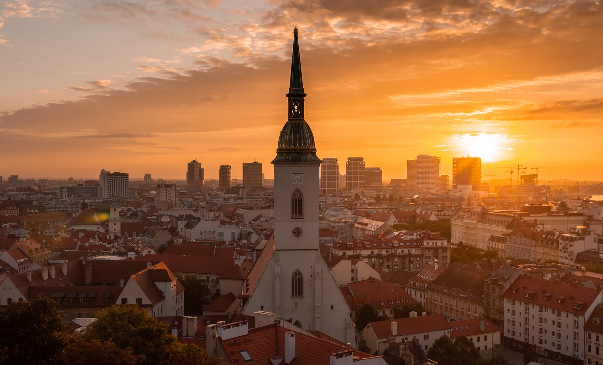 Západ slnka nad panorámou mesta s vysokou kostolnou vežou v popredí, obklopenou budovami s červenými strechami. Obloha žiari teplými oranžovými odtieňmi.